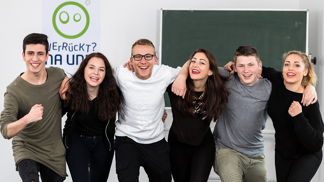 Sechs Jugendliche stehen lachend nebeneinander vor einer Schultafel und dem Verrückt-Na-und-Logo